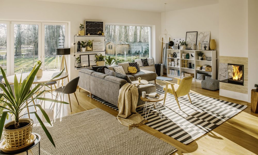 Deux tapis au sol dans un intérieur de salon scandinave blanc avec grande fenêtre, porte vitrée, canapé d’angle, cheminée et plantes fraîches
